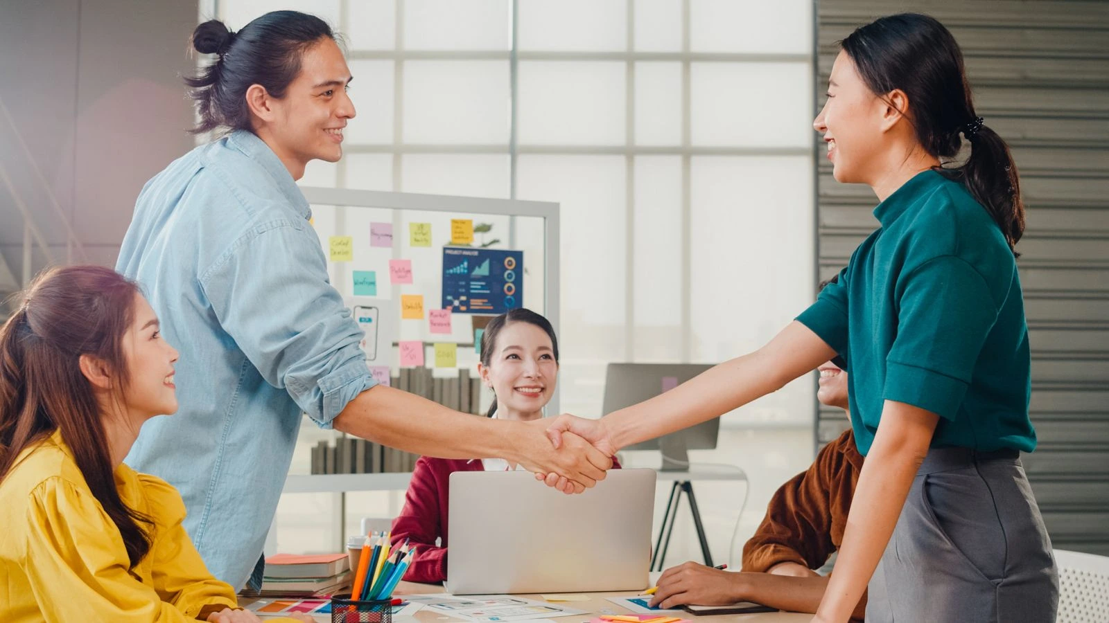 Dua rekan kerja berjabat tangan di depan tim saat rapat di kantor